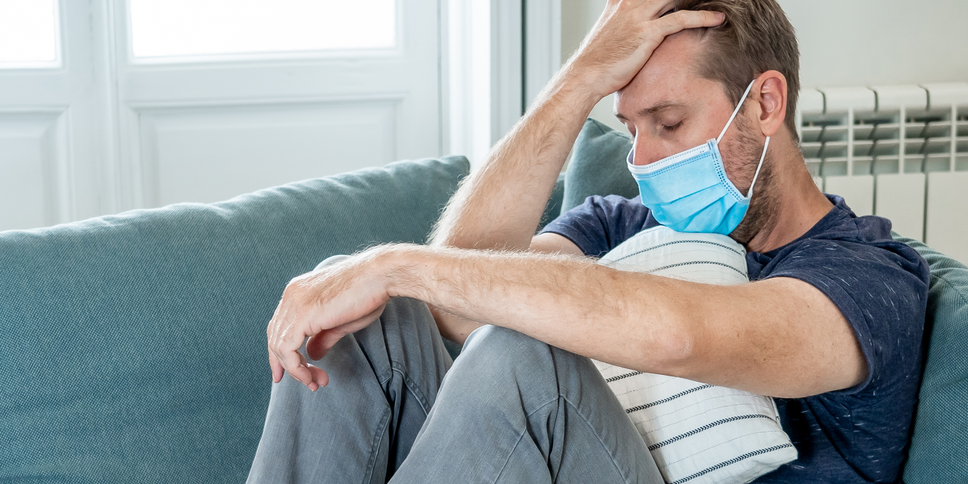 man with mask on sofa hugging pillow, hand on head